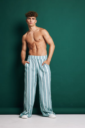 Young man with curly hair posing shirtless in striped wide pants and white sneakers against green background. Fashion model standing with hands in pockets and confident expression.の写真素材