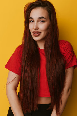 Woman with long straight hair wearing red crop top posing against yellow background smiling confidently. Portrait of young female with natural makeup and casual style in studio.の写真素材