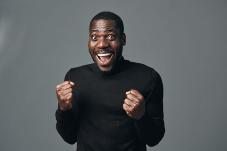 African man with dark skin wearing black sweater showing excitement and joy with clenched fists and wide smile in creative conceptual visual style studio portrait.の写真素材