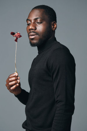 African man with dark skin holding dried red flower in hand wearing black turtleneck sweater against gray background in creative visual designer style portrait.の写真素材