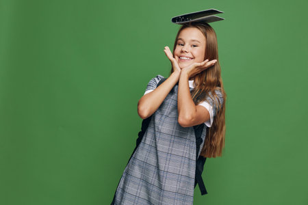 Schoolgirl smiling with hands near face and notebook on head wearing backpack standing in front of green background. Child with long hair and uniform for education concept.の写真素材