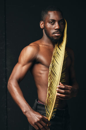 Black man with muscular body holding golden palm leaf in creative conceptual visual style. African ethnicity, artistic portrait with dark background and strong lighting for contrast.の写真素材