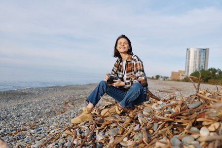 Beach photographer, woman sitting with camera on a pebbled shore, casual plaid shirt and jeans, sunny coastal day, relaxed pose capturing candid moments and expansive ocean sceneryの写真素材