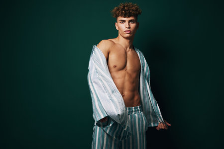 Young man with curly hair wearing striped pajamas posing shirtless against dark background, showing toned body and confident expression in studio lighting.の写真素材