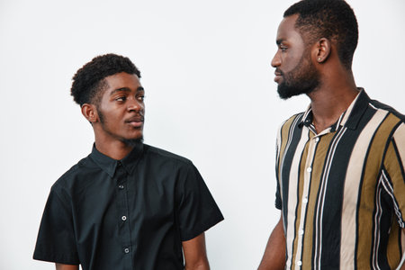 Two african american men with dark skin standing and looking at each other in casual shirts against white background. Young black males with natural hair and facial hair indoors.の写真素材