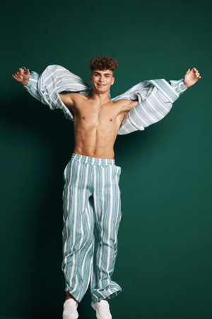 Young man with curly hair wearing striped pants and shirt jumping against green background. Casual style, smiling, energetic and confident male model in motion.の写真素材