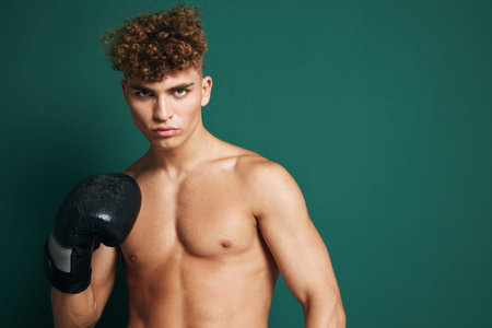 Young man with curly hair and boxing glove in training pose against dark green background expressing focus, strength and athleticism with bare skin above waist.の写真素材