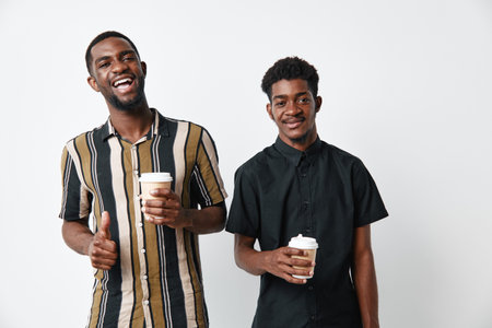 Two young black men with dark skin holding coffee cups smiling and posing on white background. Casual clothing, friendship and positive mood with beverage in hands.の写真素材