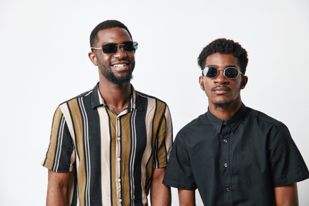 Two young black men with dark skin wearing sunglasses and casual shirts standing together on white background smiling and posing for portrait photography.の写真素材