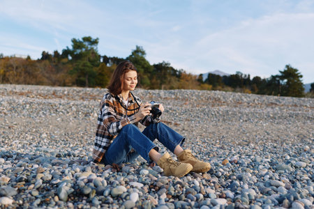 Woman photographer sits on a pebble beach, holds a camera and photographs the scenic shoreline during daylight, casual plaid shirt and boots, calm natural setting.の写真素材