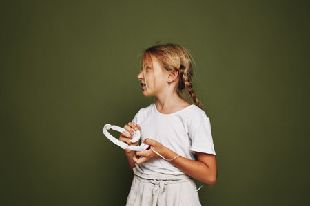 Young girl with braided hair holds white headphones and looks sideways on an olive green background. Casual outfit with a thoughtful expression. Studio portrait.の写真素材