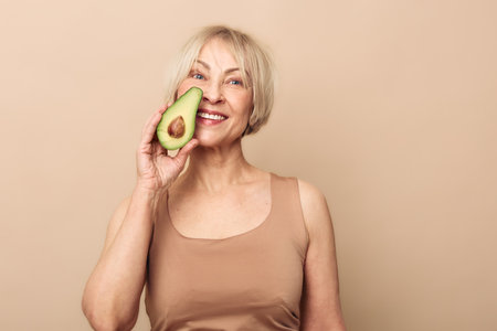 Smiling mature woman holds half of avocado near eye, posing in beige tank top on neutral background. Concept of healthy lifestyle, natural beauty, organic food, wellness and self-care.の写真素材