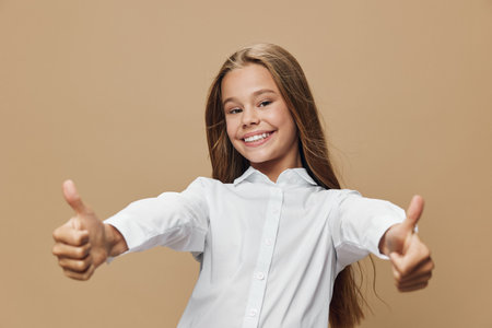 Smiling girl in white shirt showing thumbs up gesture with both hands, positive expression and casual look on beige background. Happy child with long hair and confident pose.の写真素材