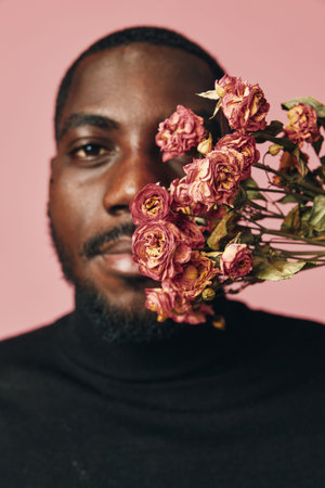 African man with dark skin holding dried pink roses close to face in creative conceptual visual style on pink background. Portrait with flowers and artistic expression in studio.の写真素材