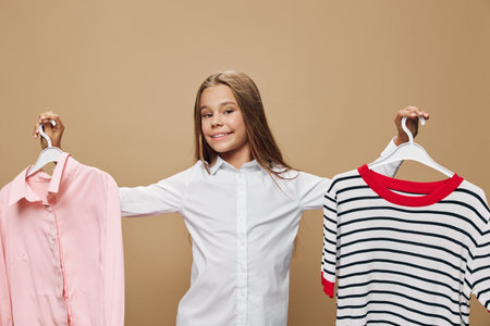 Girl with long hair smiling and holding two hangers with clothes, pink shirt and striped sweater with red collar on beige background. Young child wearing white shirt, choosing outfit.の写真素材