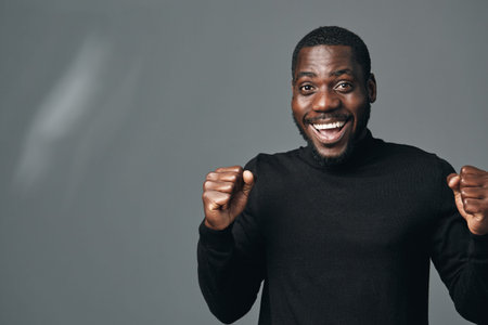 African man with dark skin wearing black sweater smiling and celebrating success with clenched fists in creative conceptual visual style on gray background.の写真素材