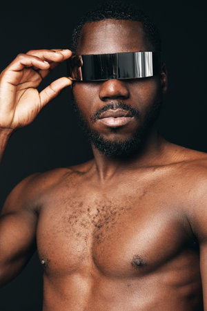 Black man with bare chest wearing futuristic silver visor in creative conceptual visual style. Portrait of African ethnicity with modern metallic eyewear on dark background for innovation and fashion.の写真素材