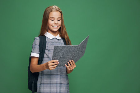 Schoolgirl with backpack reading document or folder standing against green background for education,...