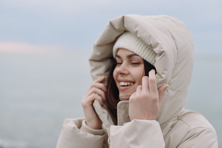 Winter, woman, outdoors, hooded jacket, smiling as she adjusts her beanie, cold breeze and soft...