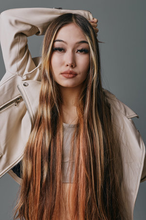 Young woman with long straight hair wearing beige jacket posing with one arm raised on neutral...