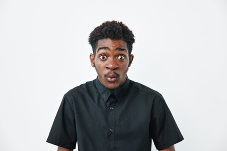 Young African American man with dark skin and curly hair wearing black shirt looking surprised with wide eyes and open mouth on white background in studio portrait.の写真素材