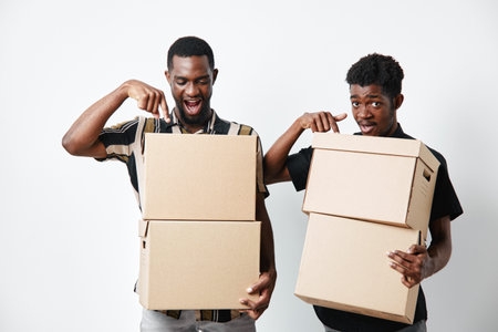 Two african american men with dark skin holding cardboard boxes for delivery and moving, smiling and pointing at packages on white background in studio.の写真素材