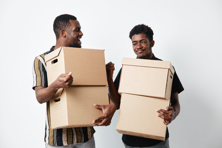 Two african american men with dark skin holding cardboard boxes for moving or delivery, smiling and standing on white background in casual clothes.の写真素材