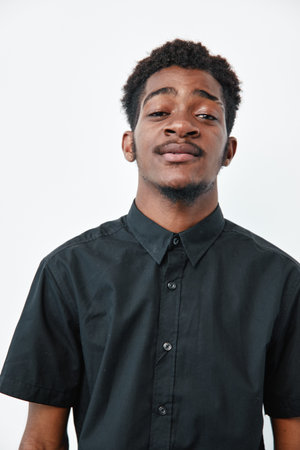 Young black man with dark skin wearing black shirt standing against white background looking confident and relaxed with slight smile and short curly hair in studio portrait.の写真素材
