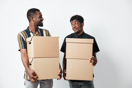 Two african american men with dark skin holding cardboard boxes for moving or delivery. Casual clothing, teamwork and cooperation in logistics or relocation activity.の写真素材
