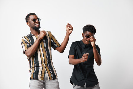 Two black men with dark skin dancing and smiling wearing sunglasses and casual shirts on white background. Friends enjoying music and having fun together in studio.の写真素材