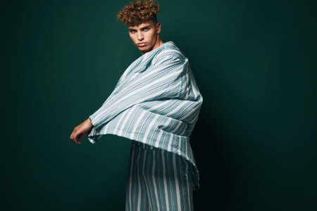 Young man with curly hair wearing striped oversized shirt and pants posing against dark green background in studio for fashion and style concept.の写真素材