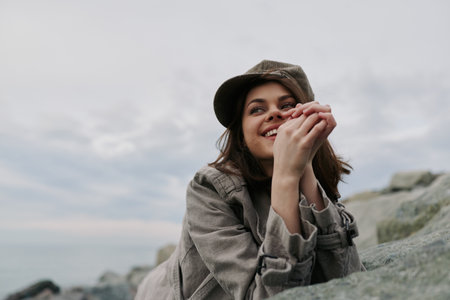 Young woman in a camouflage jacket and hat smiles while resting on rocks by the sea under an overcast sky. Concept for nature, outdoor, relaxation, and happiness.の写真素材