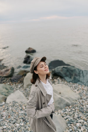 Young woman in beige coat and hat enjoys fresh air with closed eyes on rocky beach near calm sea. Concept of relaxation, peace, nature, and outdoor lifestyle at coast.の写真素材