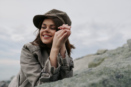 Smiling young woman in a brown plaid coat and hat leans on a rock outdoors under cloudy sky. Natural light highlights her joyful expression and casual autumn fashion style.の写真素材