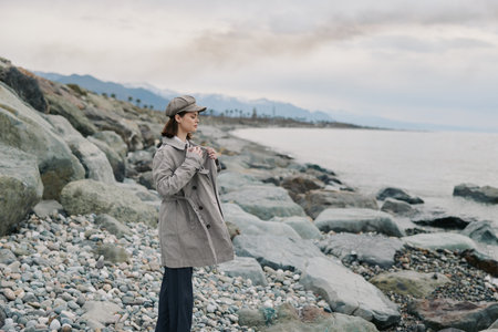 Young woman in a plaid coat and hat stands on a rocky beach looking at the sea. Cloudy sky and...