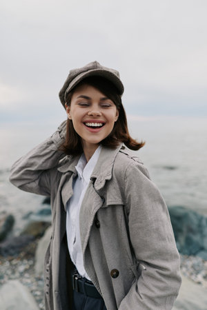 Smiling young woman in checkered coat and cap stands near rocky shore with closed eyes, enjoying fresh air and natural outdoor atmosphere by the sea on cloudy day.の写真素材
