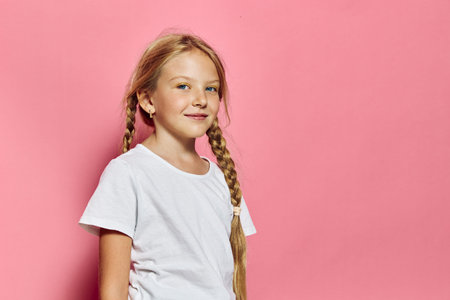 Smiling girl with braided hair wearing white t-shirt stands against pink background in a bright studio. Concept for childhood and happiness.の写真素材