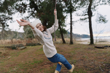 Woman on a wooden swing in a park, wearing a knit sweater and jeans, smiling as she swings, trees in the background, autumn light, carefree moment in nature, outdoor joyの写真素材