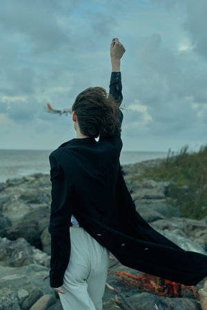 Victory stance on a rocky coast as a figure raises a fist, cape fluttering, ocean wind and dramatic silhouette convey triumph, empowerment, and bold determination.の写真素材