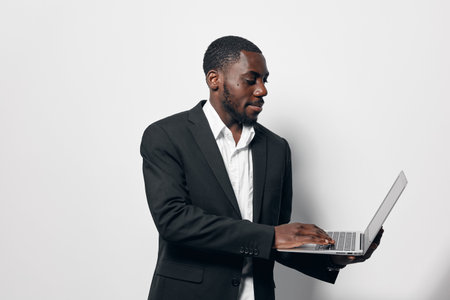 Black man with dark skin wearing black suit and white shirt working on laptop standing against white background. Business professional focused on technology and communication.の写真素材