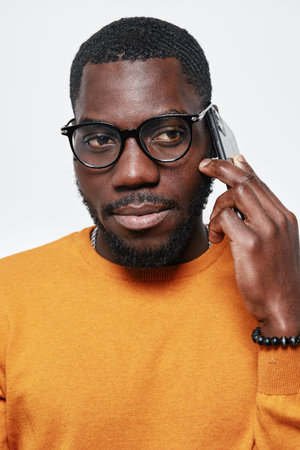 African American man with dark skin wearing glasses and orange sweater talking on smartphone, closeup portrait with serious expression on white background.の写真素材