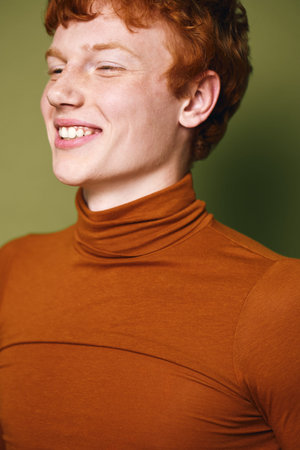 Young man with red hair wearing orange turtleneck smiling with eyes closed in studio. Casual...
