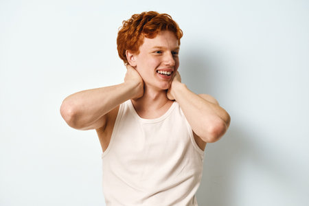 Young man with red hair and freckles in white tank top smiling and touching neck in casual studio portrait with white background and natural expression.の写真素材