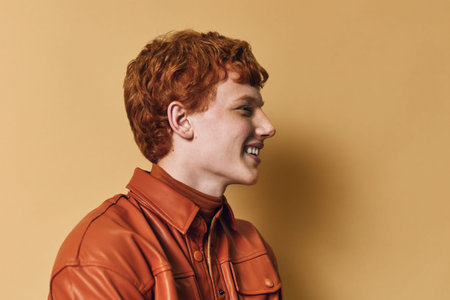 Redhair young man in orange leather jacket smiling showing profile on yellow background. Portrait of happy casual male model with curly hair and bright expression wearing stylish outfit.の写真素材