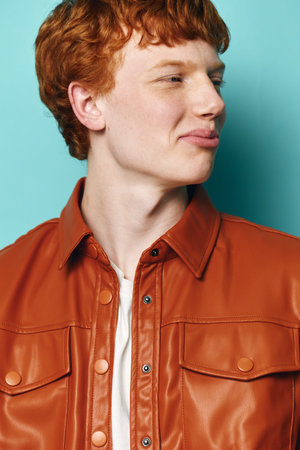Young man with red hair wearing brown leather jacket and white shirt posing in profile against plain turquoise background with casual expression and natural light.の写真素材