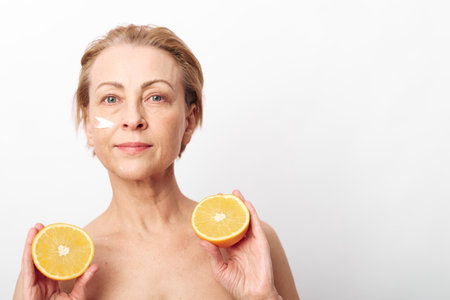 Mature woman holds two halves of an orange while applying cream on her face. Concept of natural skincare, healthy aging, vitamin care, and organic beauty treatment in bright studio setting.の写真素材