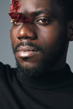 African man with dark skin and beard in creative conceptual style closeup portrait with dried red flower on face wearing black turtleneck against gray background.の写真素材