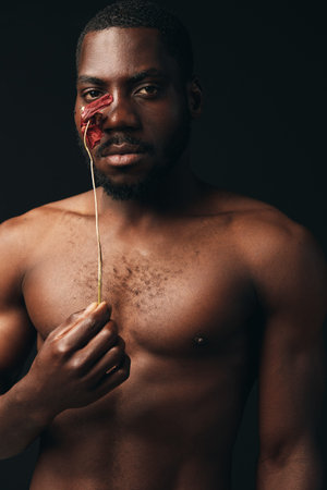 Black man with bare chest holding dried flower near face in creative conceptual visual style. Portrait of African ethnicity with artistic expression on dark background for modern art.の写真素材