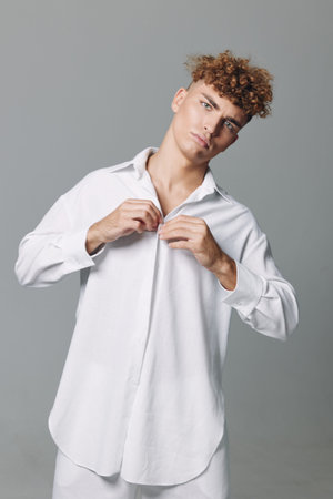 Young man with curly hair wearing oversized white shirt buttoning it up against plain gray background. Casual fashion, studio portrait, confident expression and modern style for lifestyle.の写真素材