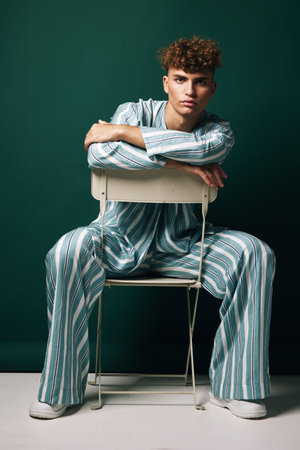 Young man with curly hair wearing striped pajamas sitting on chair backwards against green...
